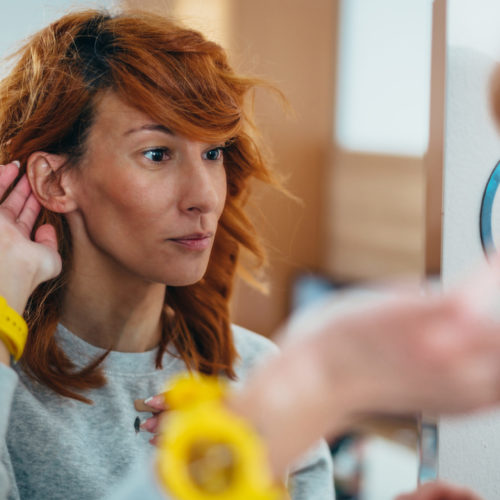 Young redhead woman putting a hearing aid in her ear while looking herself in the mirror